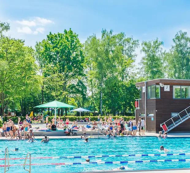 A vibrant blue sky complements a bustling pool area, with individuals swimming and trees lining the edge for shade.